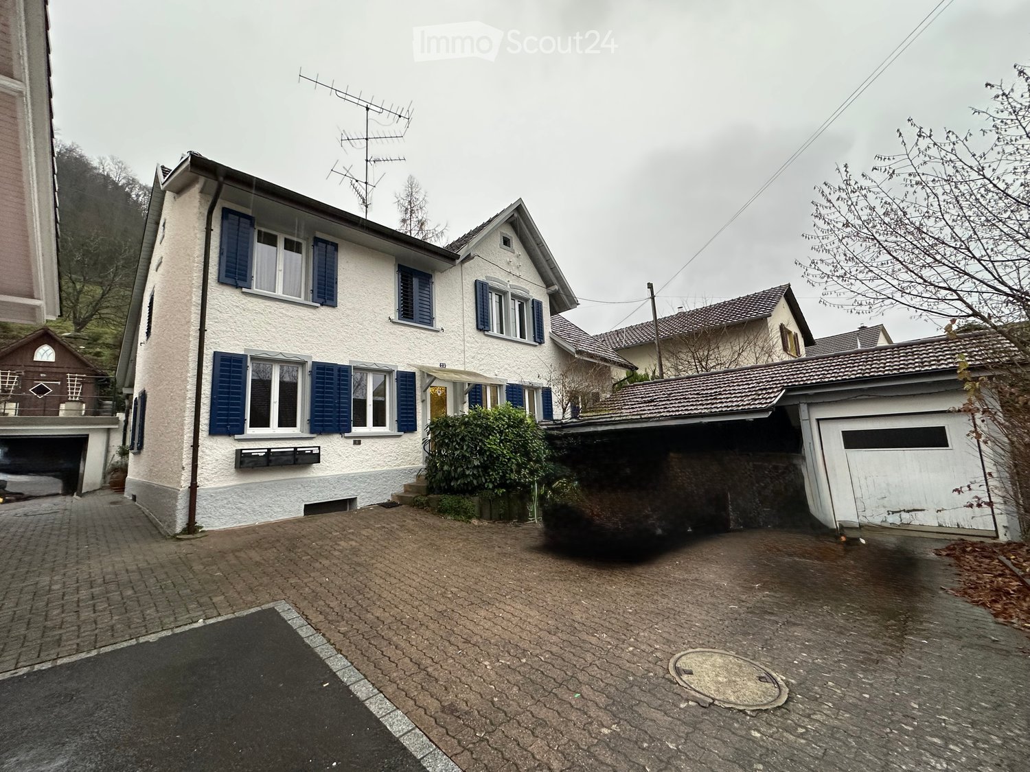 Two-story house with white exterior, blue shutters, and a garage. The driveway is paved, and there are some bushes near the entrance.