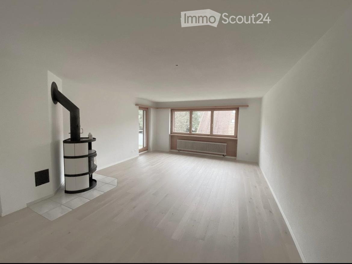 Empty room with light wooden flooring, white walls, large windows, and a built-in radiator. A black chimney and a small black cylindrical object are visible on the left side.