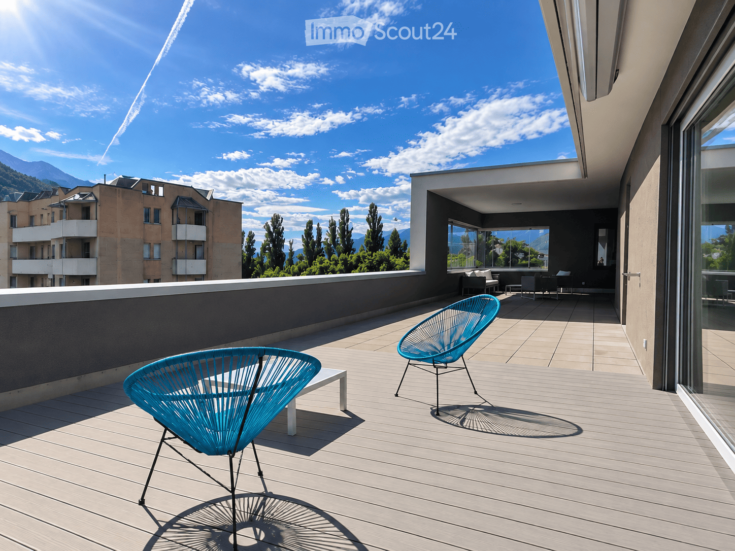 Modern rooftop terrace with wooden decking, two turquoise chairs, and a view of mountains and cityscape.