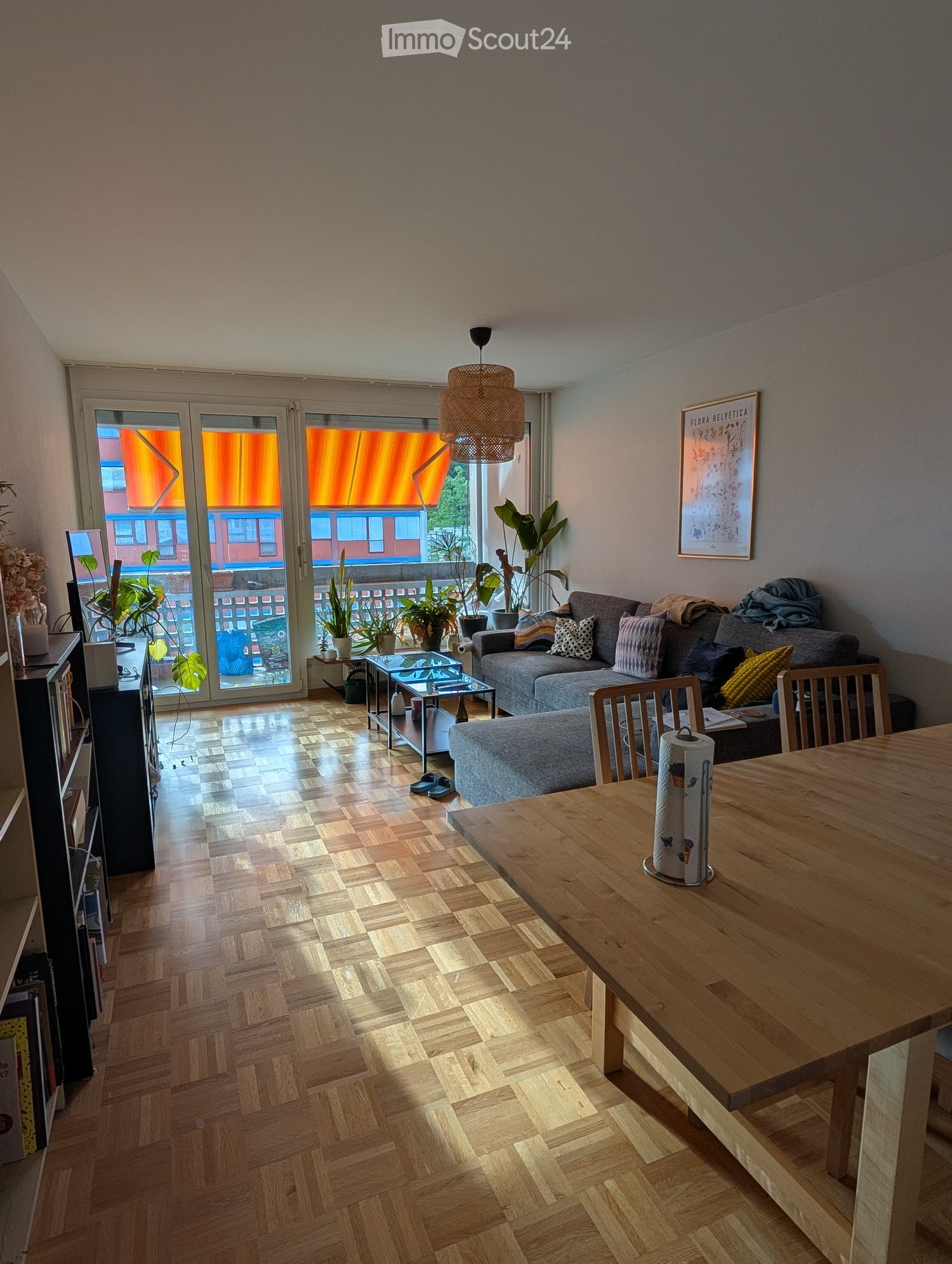 Bright living room with wooden flooring, large glass doors leading to a balcony, sofa, coffee table, dining table, and potted plants.