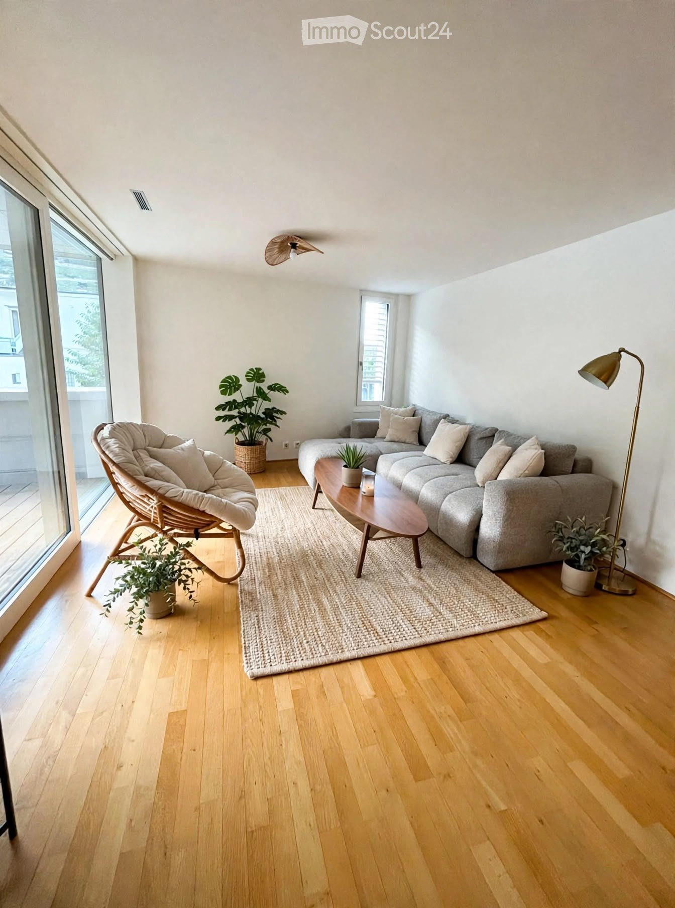 Modern living room with light wooden flooring, a grey sectional sofa, a wicker hanging chair, a wooden coffee table, a floor lamp, and several potted plants.