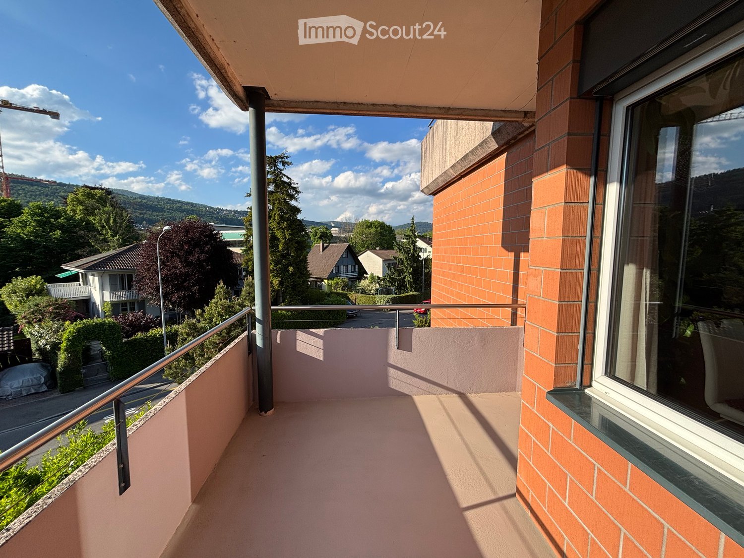 Balcony with metal railing, pink floor, overlooking a residential area with houses, trees, and a mountain in the background.