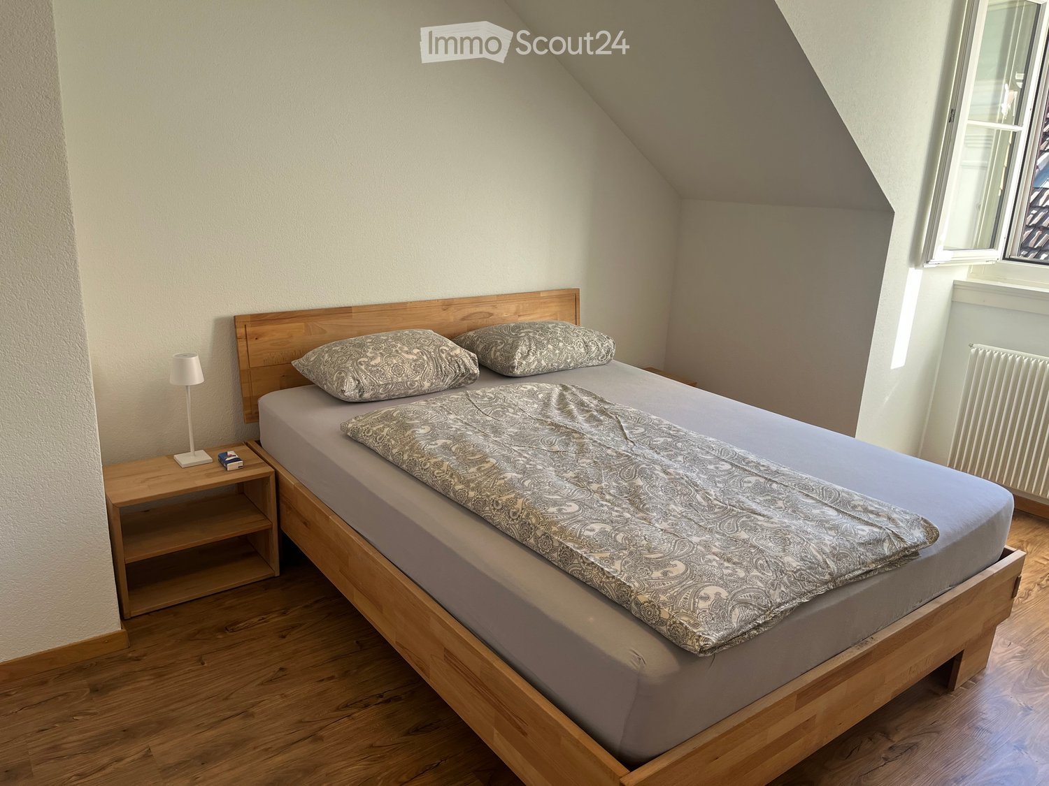 Cozy attic bedroom with wooden bed, nightstand, lamp, and radiator. The bed has a grey duvet and patterned pillows.