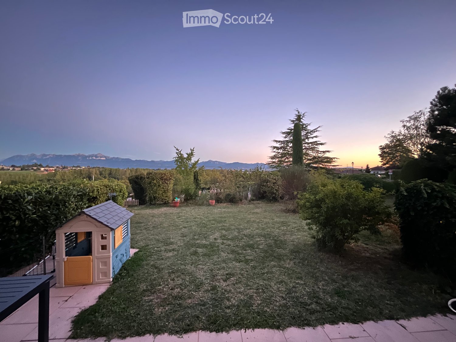 A well-maintained garden with a small playhouse, surrounded by hedges and trees, with a scenic mountain view in the background.