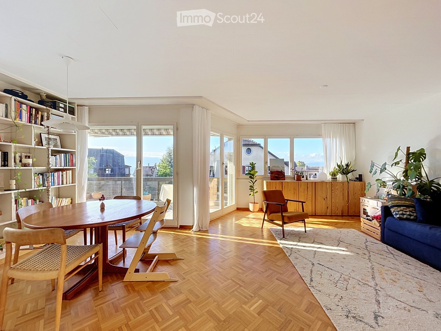 Bright, open living and dining area with wooden floors, large windows, and a view. Features a blue sofa, wooden dining table with chairs, bookshelf, and potted plants.