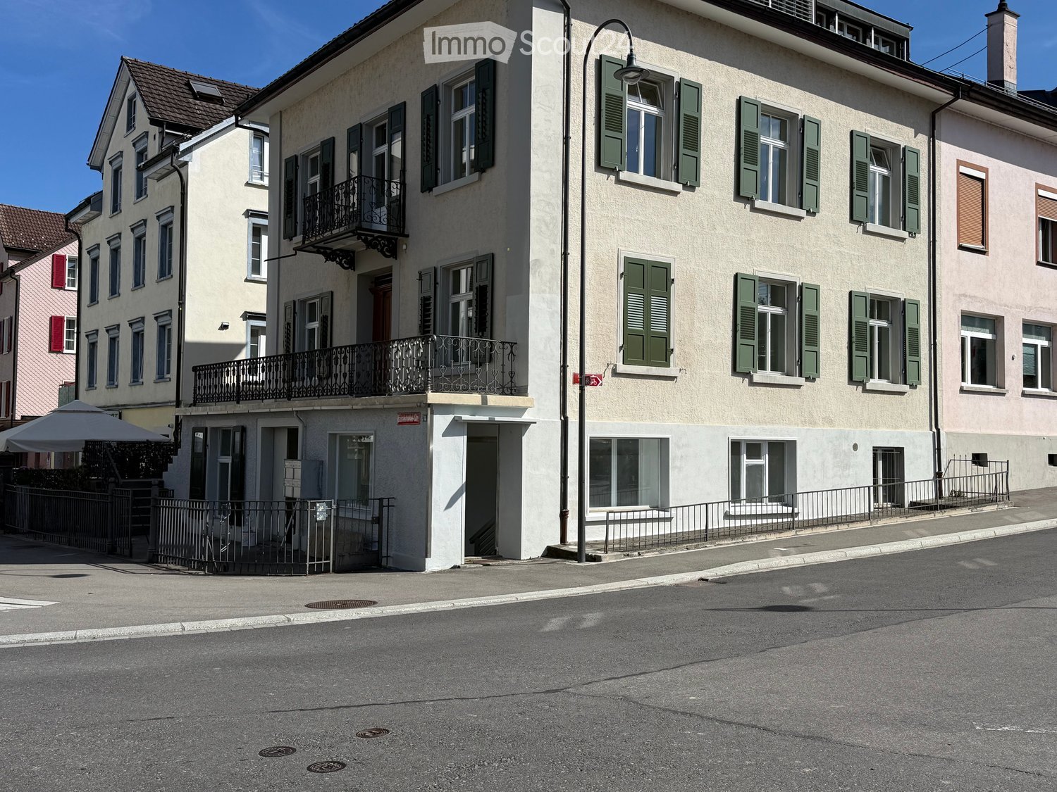Multi-story residential building with light-colored facade, green shutters, and balconies. Adjacent buildings visible with similar architectural style.