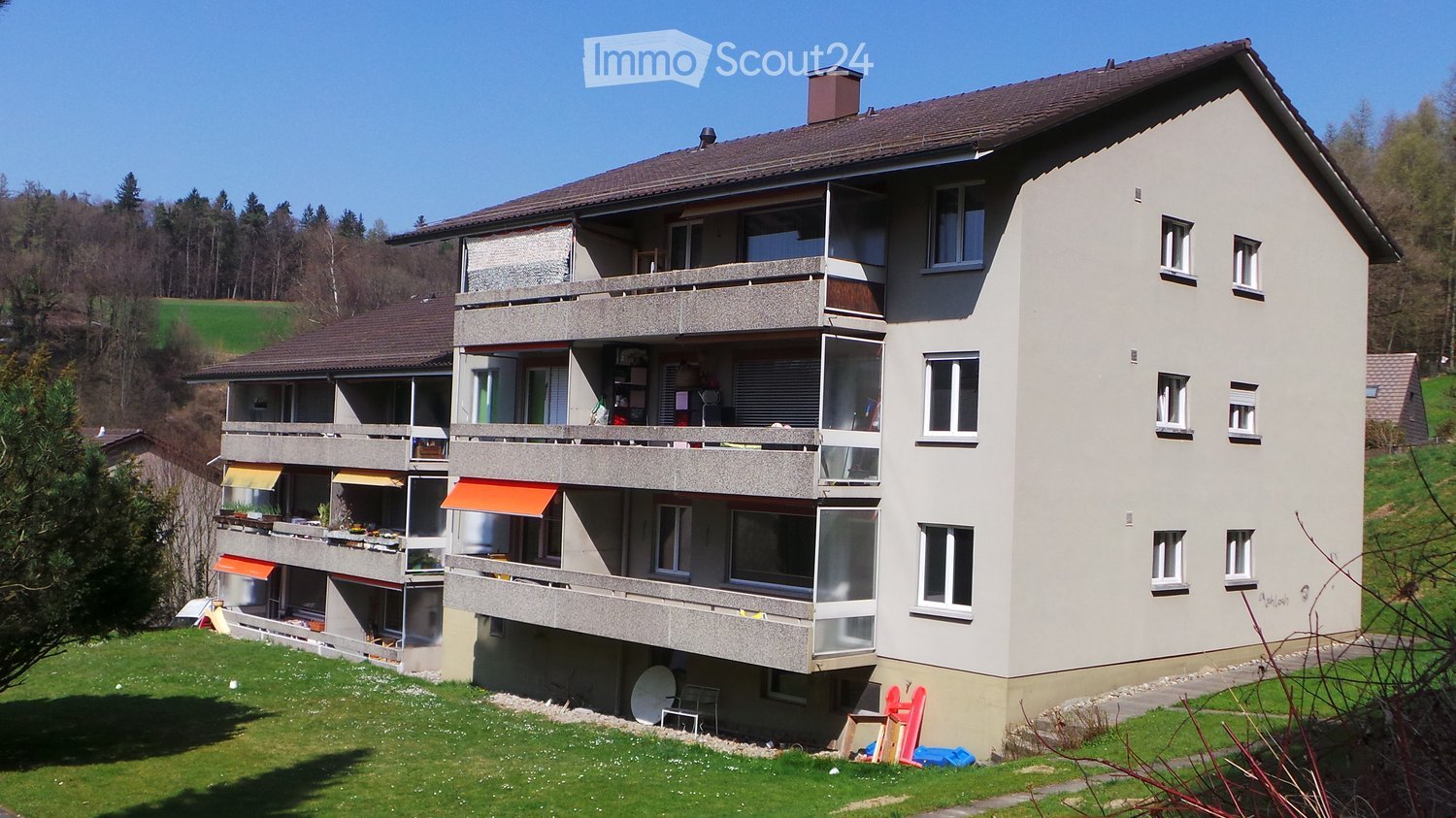 Multi-story residential building with balconies, large windows, and a surrounding green lawn.