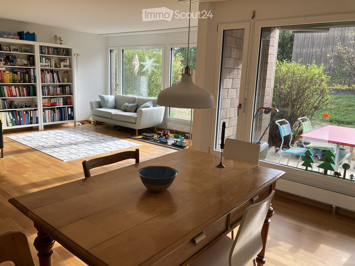 Bright living and dining area with wooden flooring, large windows, and a view of the garden. Features a grey sofa, wooden dining table with chairs, bookshelf, and a hanging pendant lamp.