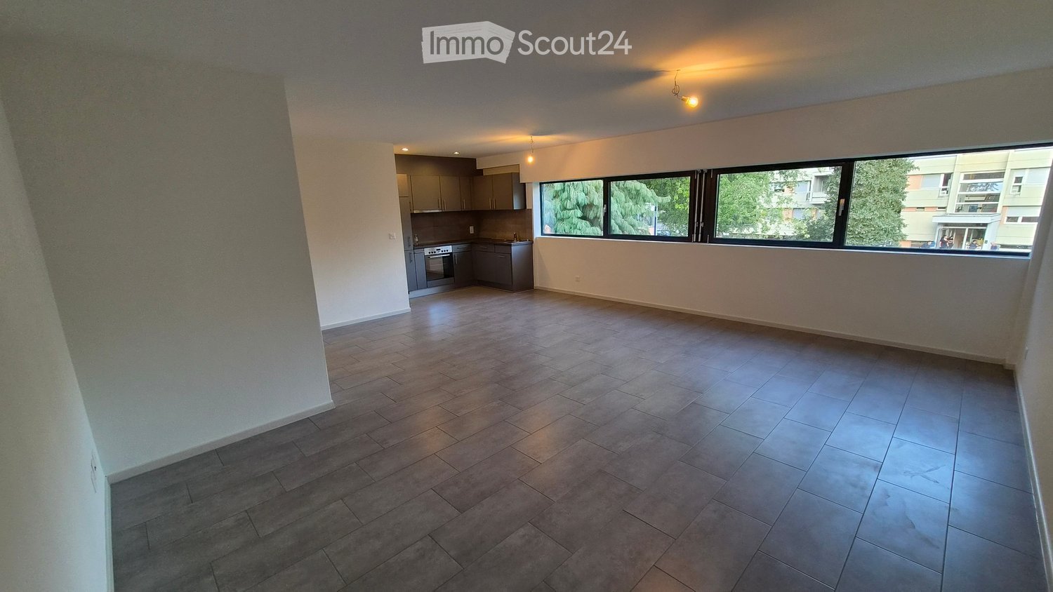 Empty room with tiled floor, large windows, and a small kitchen area with cabinets and appliances.