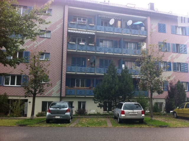 Multi-story apartment building with balconies, satellite dishes, and a parking area with several cars.