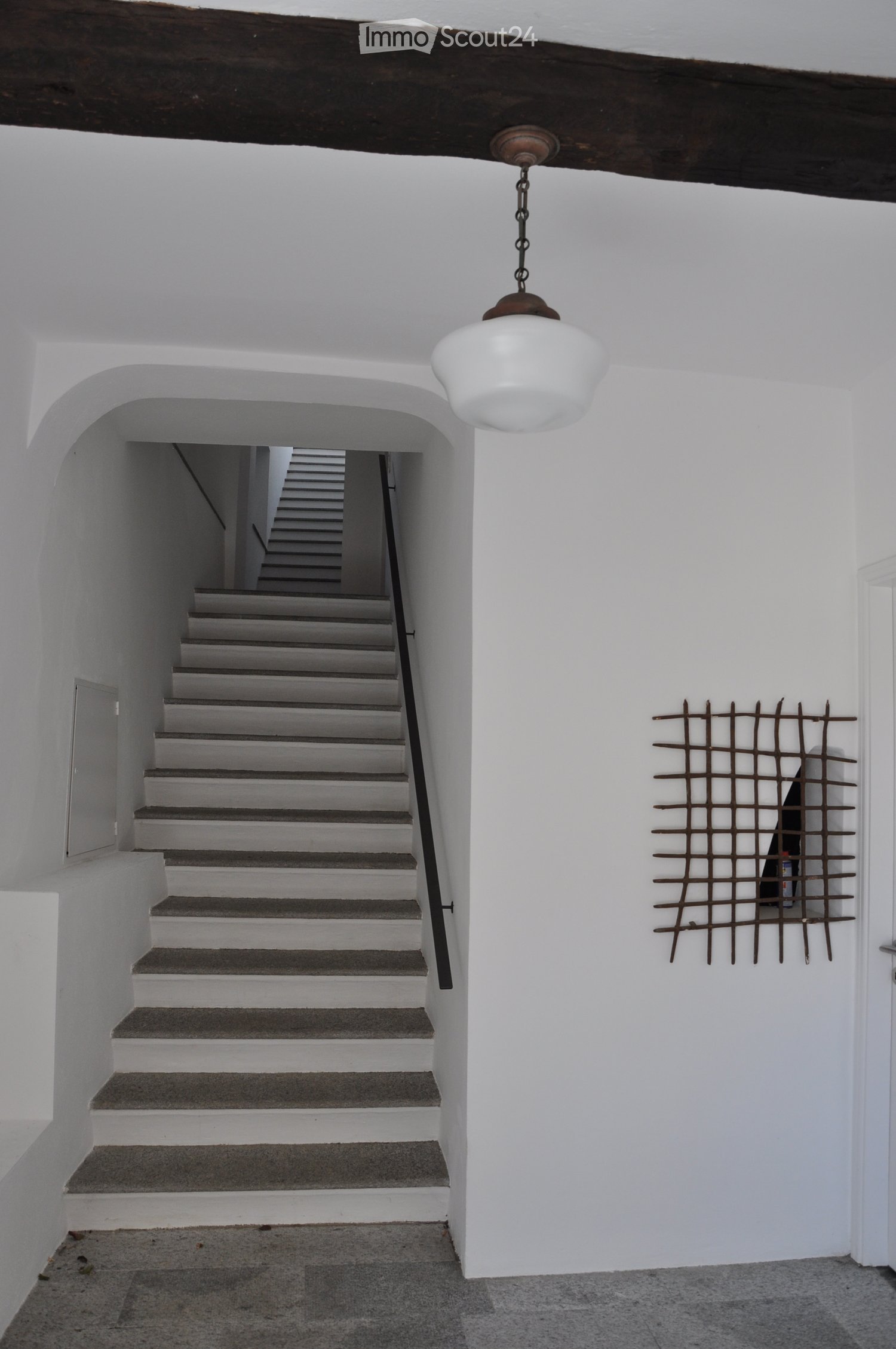A narrow hallway with white walls, a staircase leading upwards, and a decorative metal grid on the wall.