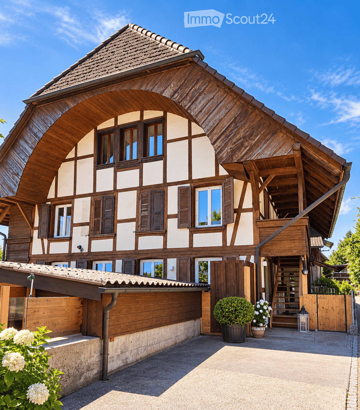 Traditional wooden house with a steeply pitched roof, white and brown facade, multiple windows with shutters, and a covered entrance area with steps leading up to the door.