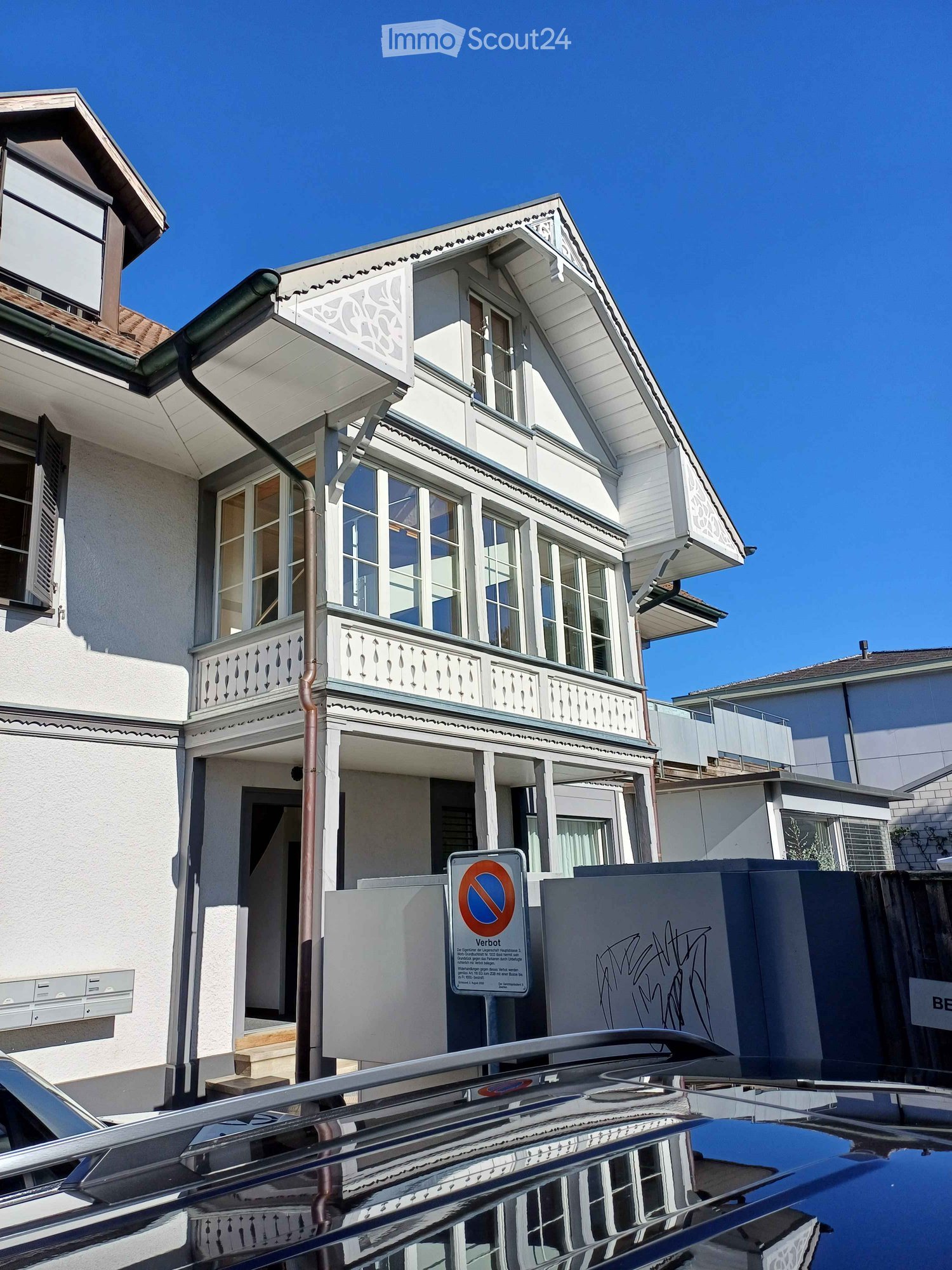 Two-story house with white exterior, multiple windows, decorative balcony, and a no-parking sign in front.