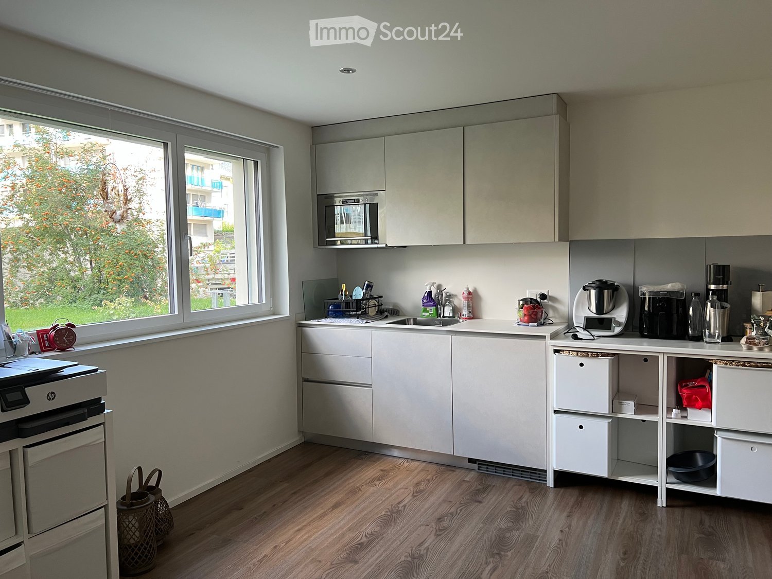 Modern kitchen with white cabinets, stainless steel appliances, wooden flooring, and a large window overlooking a green area.