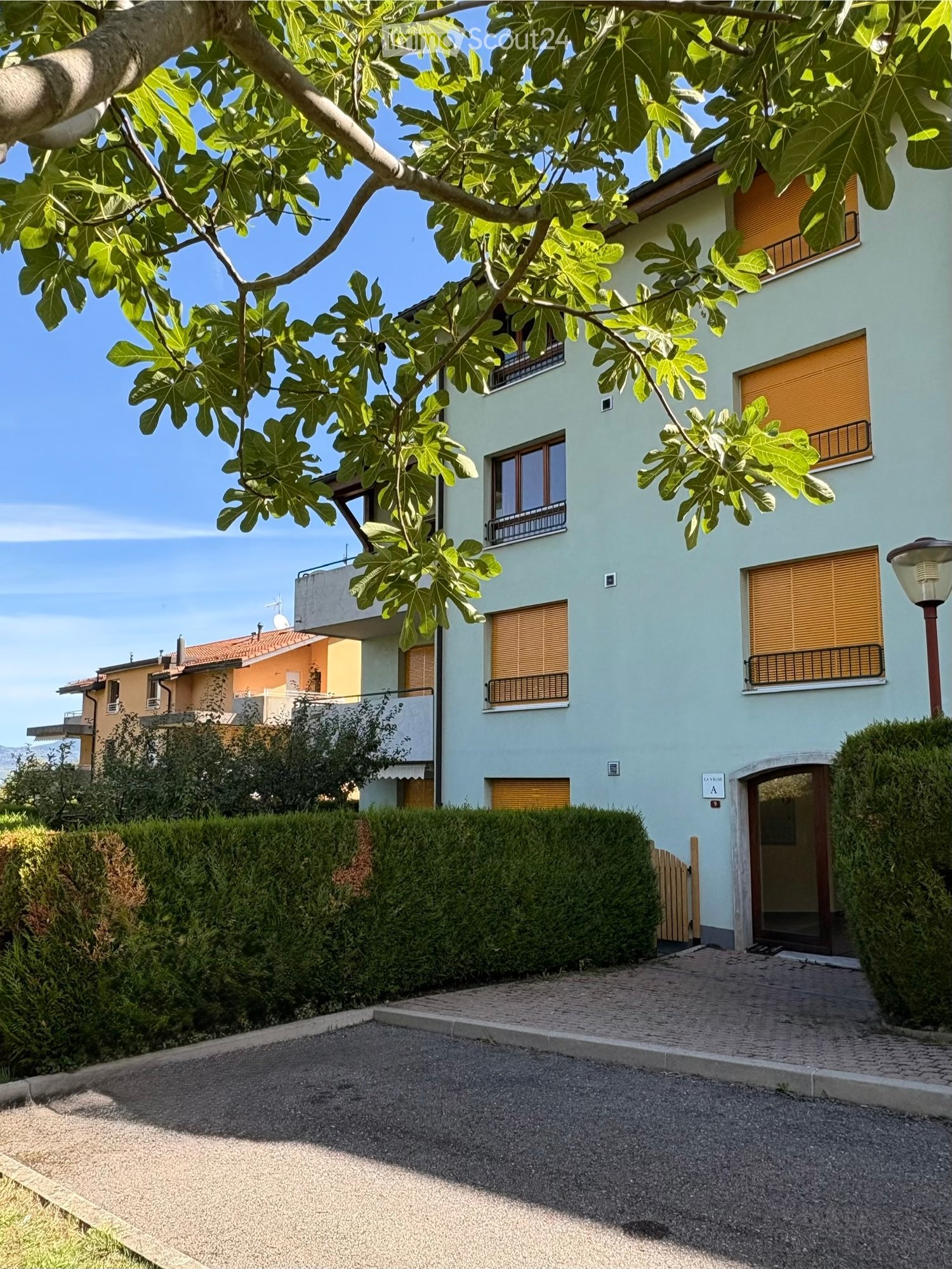 Two-story house with light blue exterior, multiple windows with shutters, and a well-maintained hedge in front.
