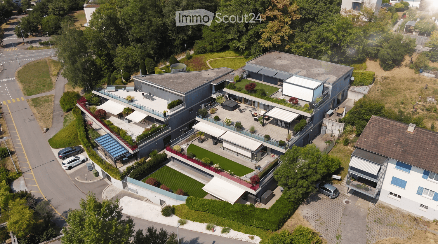 Aerial view of a modern, multi-story residential building with a spacious outdoor area featuring a lawn, patio, and seating arrangements. The building has a sleek design with large windows and a flat roof, surrounded by greenery and neighboring houses.