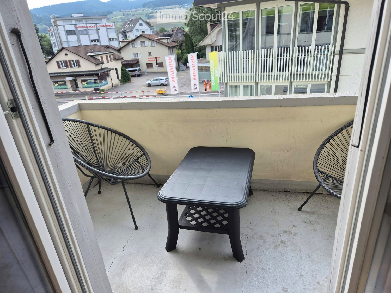 Balcony with a small black table and two black wire chairs, overlooking a town with buildings and a road.