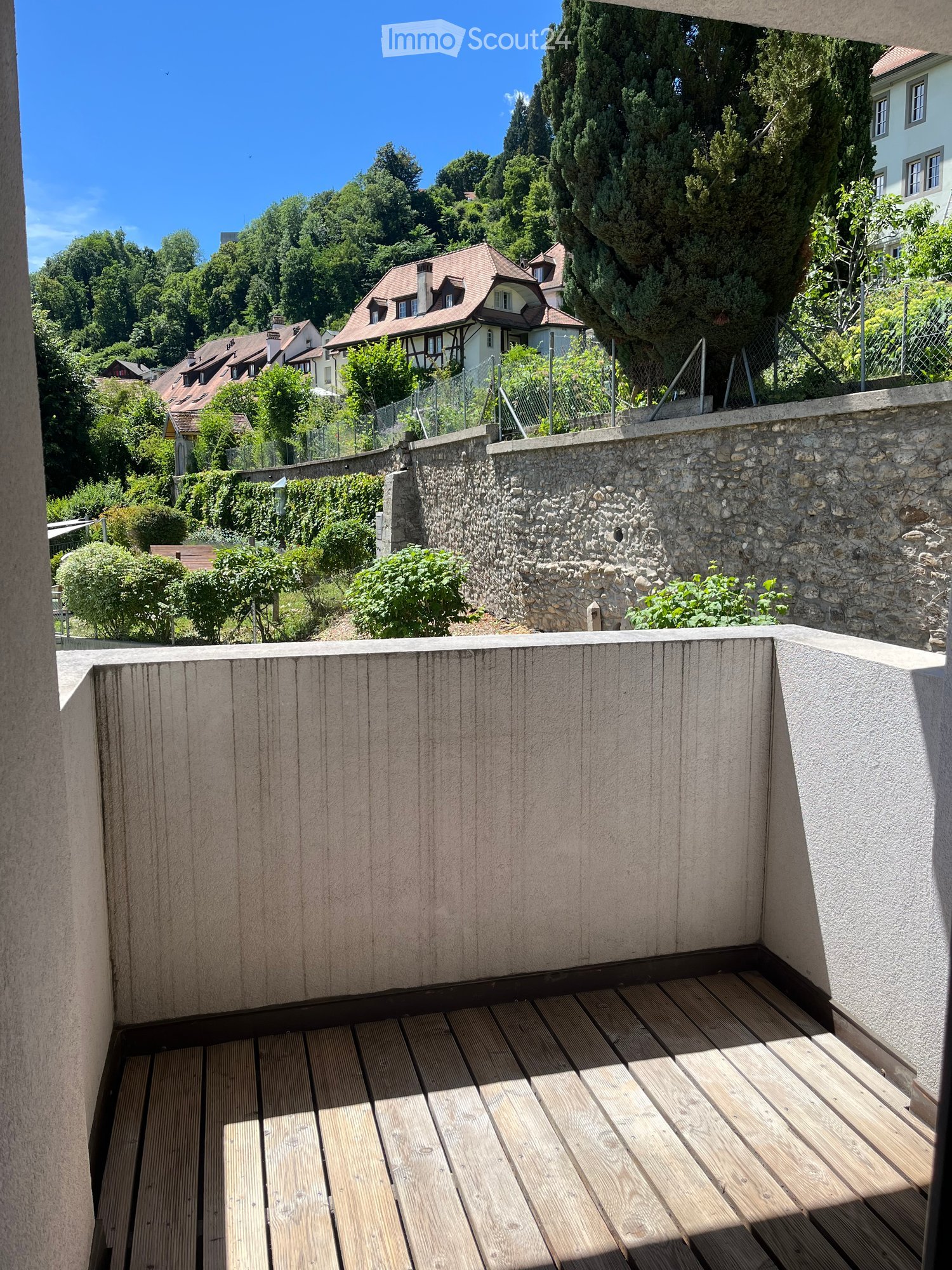 Wooden decked balcony with stone walls, overlooking a residential area with trees and houses.