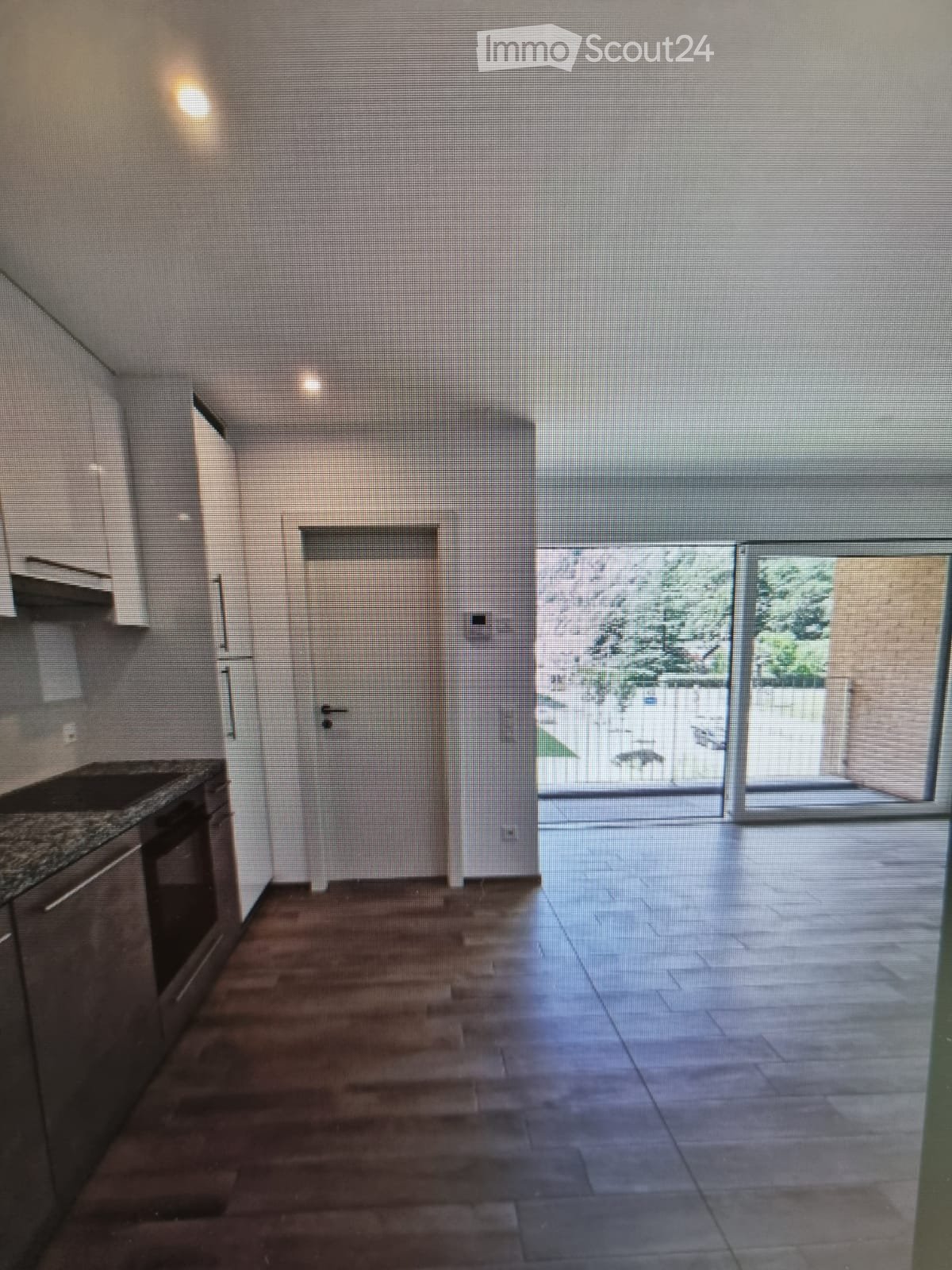 Empty room with wooden flooring, white walls, and a sliding glass door leading to an outdoor area. Kitchen area visible with cabinets and countertop.