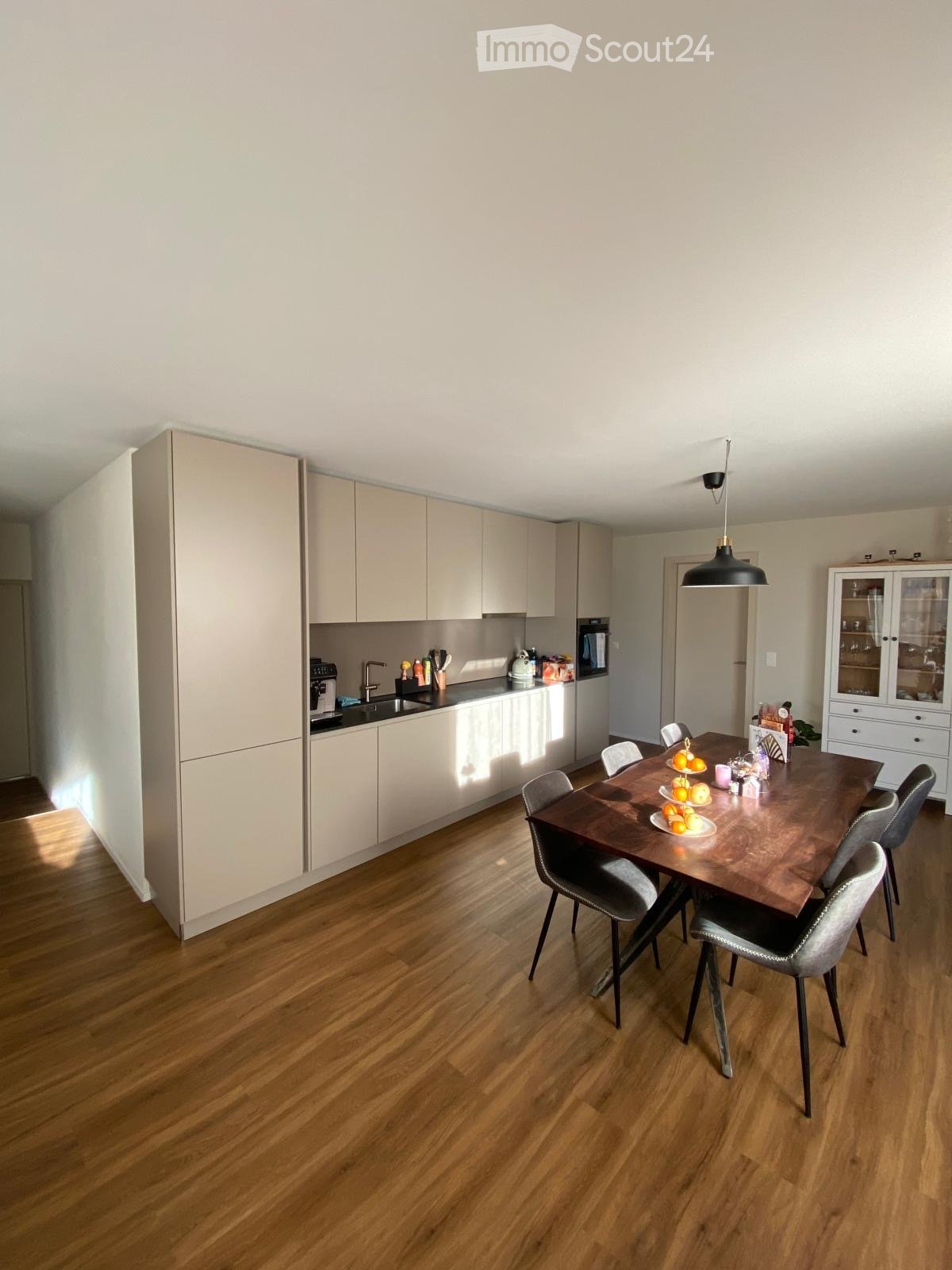 Modern kitchen with light-colored cabinets, a wooden dining table, and six chairs. The kitchen features a sink, faucet, oven, refrigerator, and dishwasher. A cabinet with glass doors is visible, and the floor is made of polished wood.