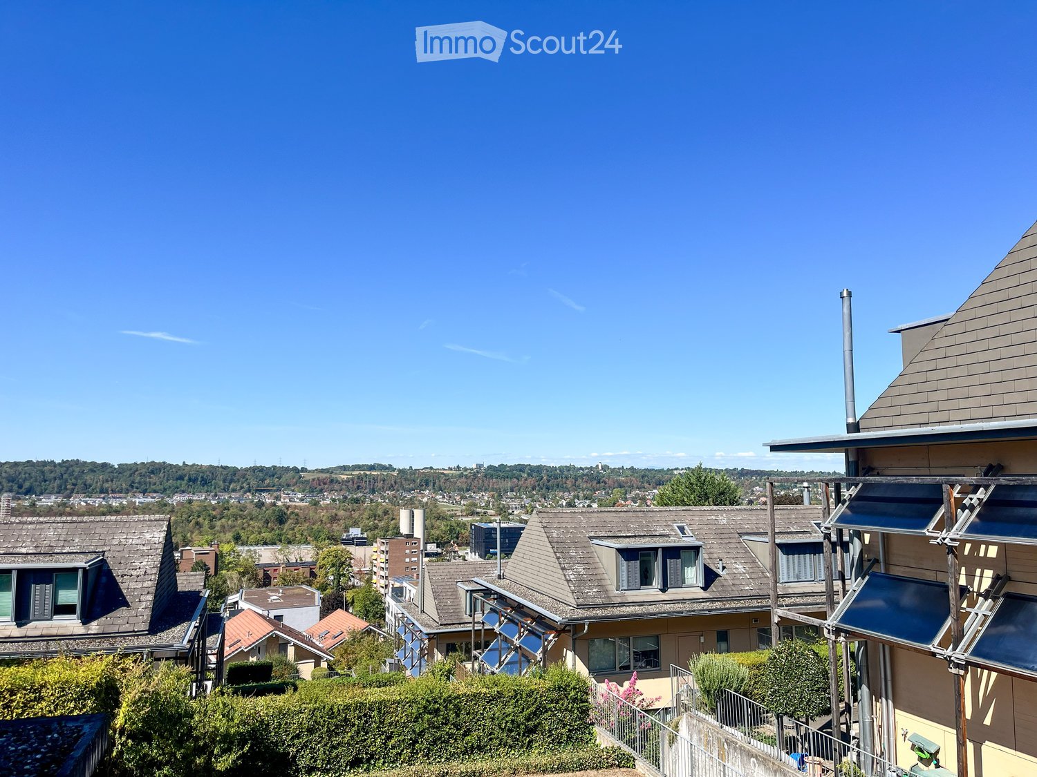 Balcony overlooking a residential neighborhood with houses, trees, and distant cityscape under a clear blue sky.