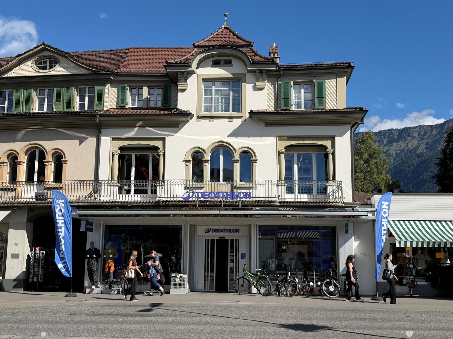 Decathlon store with a large glass front, bicycles parked outside, and a balcony with a railing above. The building has a classic European architectural style with arched windows and green shutters.