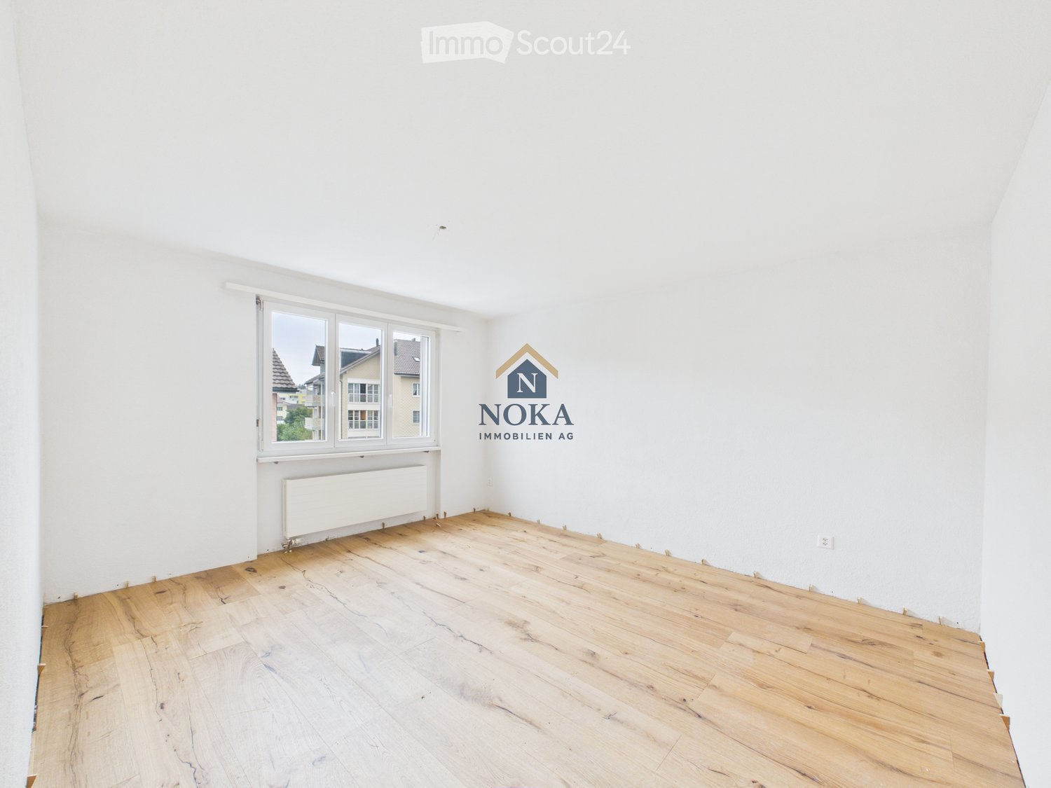 Empty room with wooden flooring, white walls, and a large window showing a view of neighboring buildings.