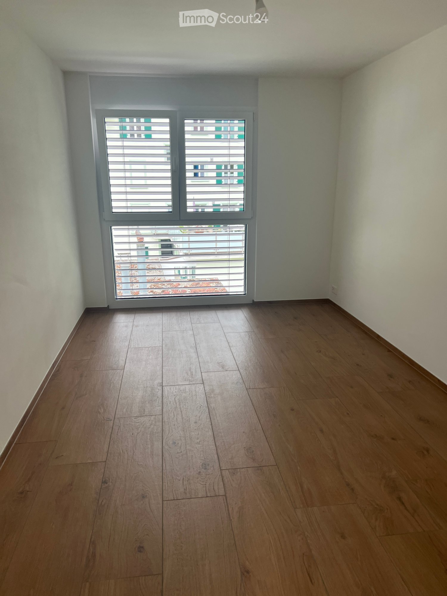 Empty room with wooden flooring and large windows with blinds, showing a view of a building exterior.