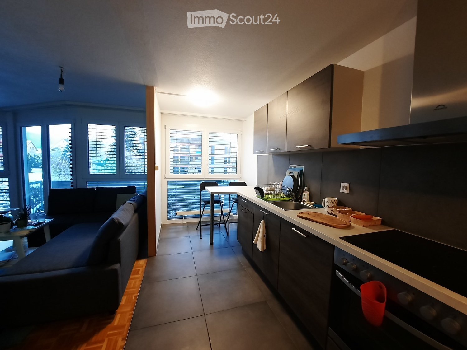 Modern open-plan living and kitchen area with grey cabinets, a sofa, a dining table with stools, and various kitchen appliances.