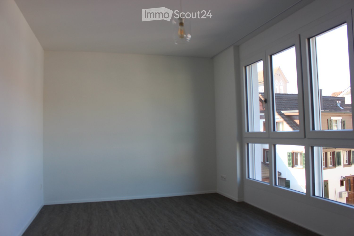 Empty room with light-colored walls, dark wooden flooring, and large windows offering a view of neighboring buildings.