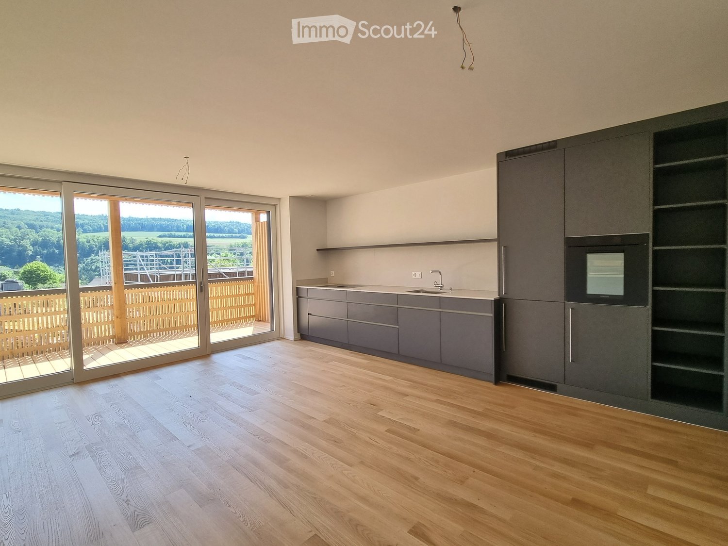 Modern kitchen with grey cabinets, wooden flooring, and large sliding glass doors leading to a balcony with a scenic view.