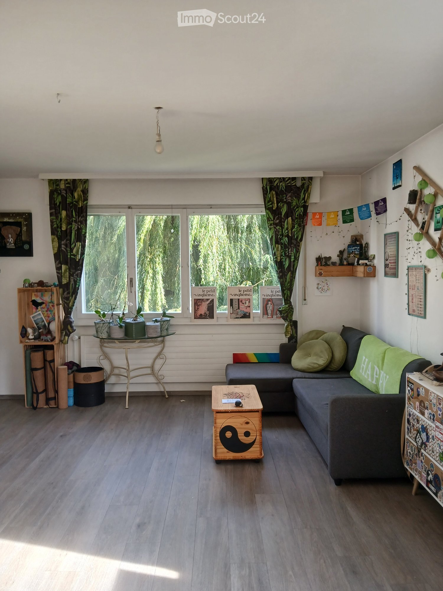 Bright living room with wooden flooring, large windows with green curtains, a grey sectional sofa with green cushions, a small wooden coffee table with a yin-yang design, a glass side table with potted plants, and shelves with decorative items.