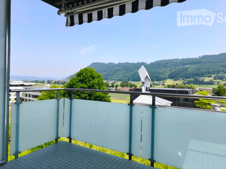 Balcony with glass railings, overlooking a scenic countryside with trees and hills under a clear blue sky.