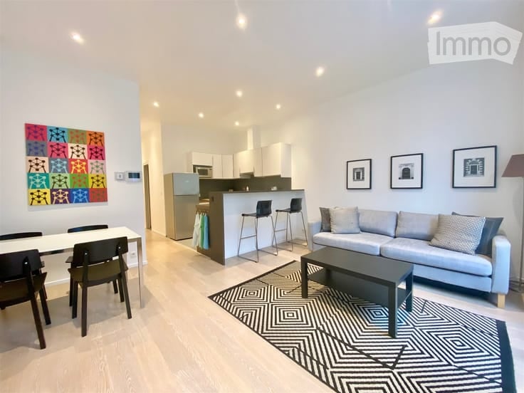 Modern open-plan living and dining area with a grey sofa, coffee table, dining table with chairs, and a kitchen area featuring a refrigerator, microwave, and bar stools. The space includes a colorful abstract painting and framed artwork on the walls, comp