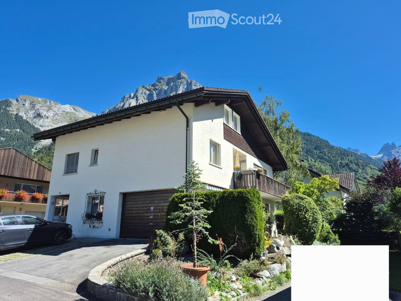 Two-story white house with a wooden garage, multiple windows, and a balcony, surrounded by a well-maintained garden and set against a backdrop of mountains.