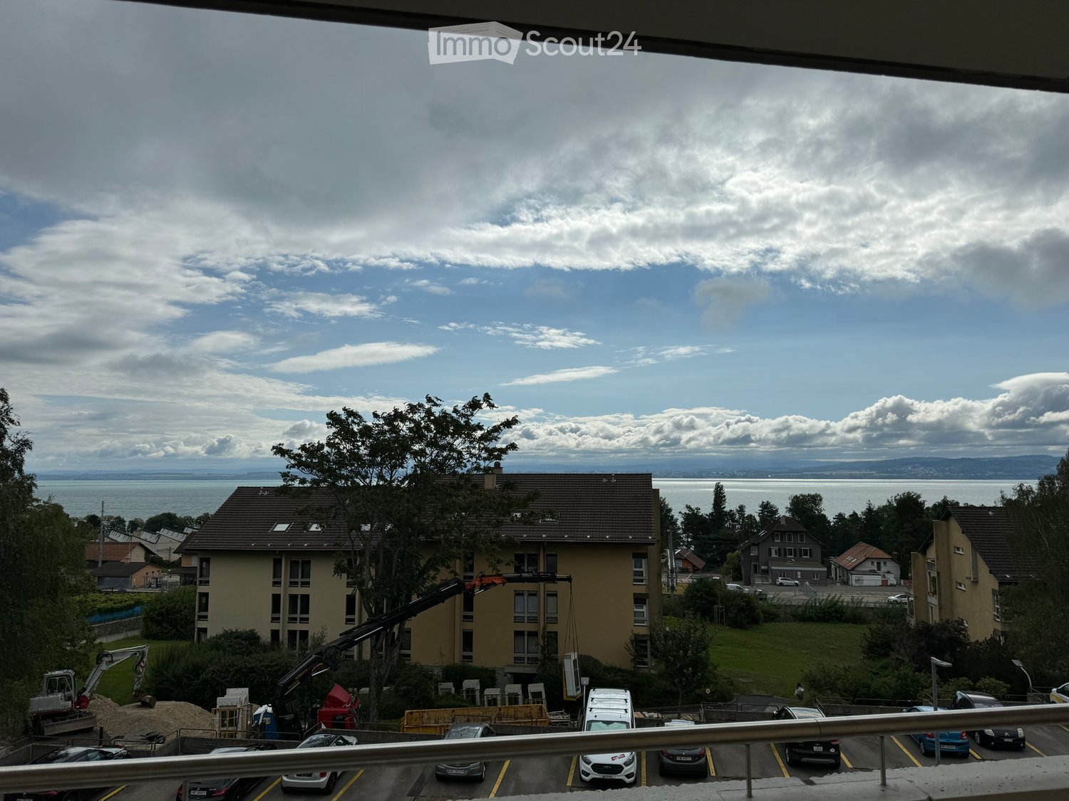 Balcony view overlooking a parking lot, several parked cars, and a residential area with buildings and a body of water in the distance.