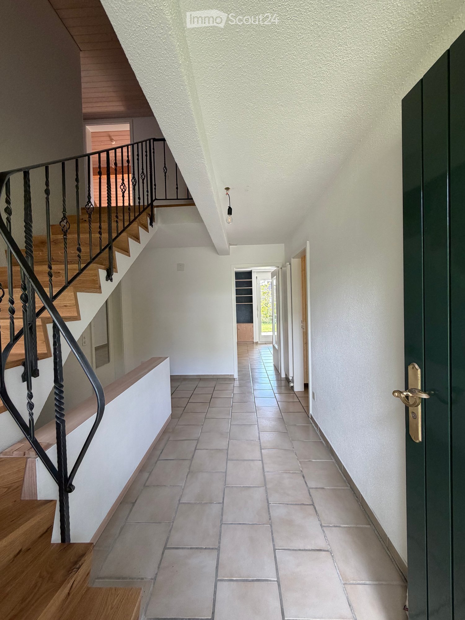 Spacious hallway with tiled floor, white walls, wooden staircase with black wrought-iron railing, and a green door at the end.