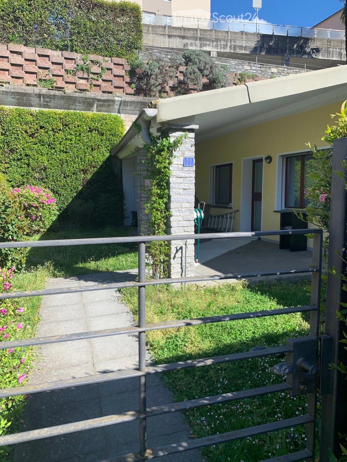 A small garden area with a pathway leading to a yellow house, featuring a metal railing, greenery, and a bench.