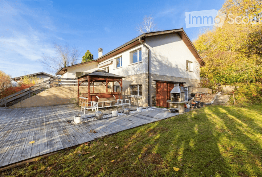 A modern two-story house with a wooden deck, outdoor seating area, and a gazebo. The garden features a well-maintained lawn, potted plants, and a barbecue area.