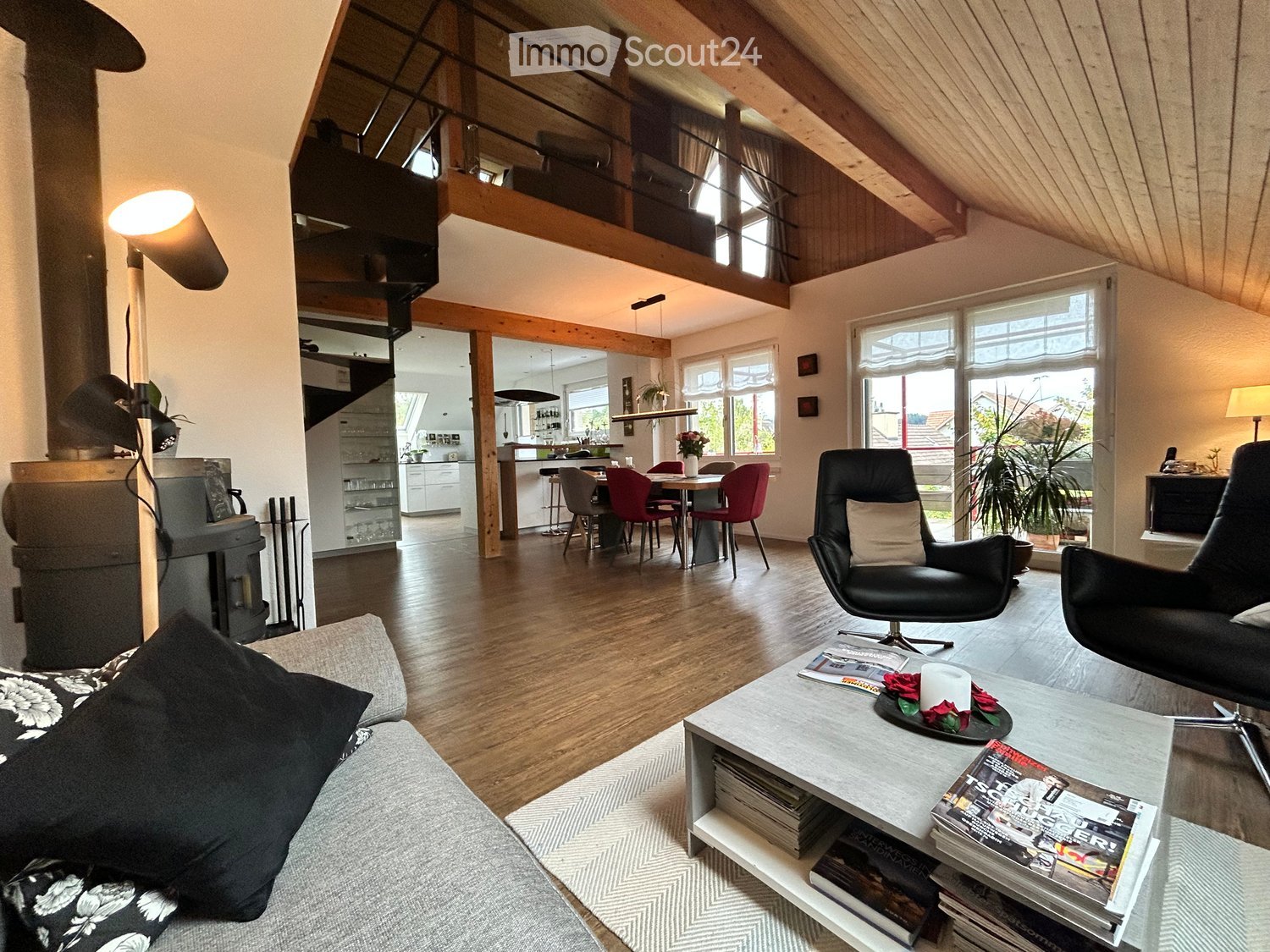 Open-plan living and dining area with wooden floors, exposed wooden beams, large windows, a sofa, coffee table, armchairs, dining table with chairs, and a fireplace.