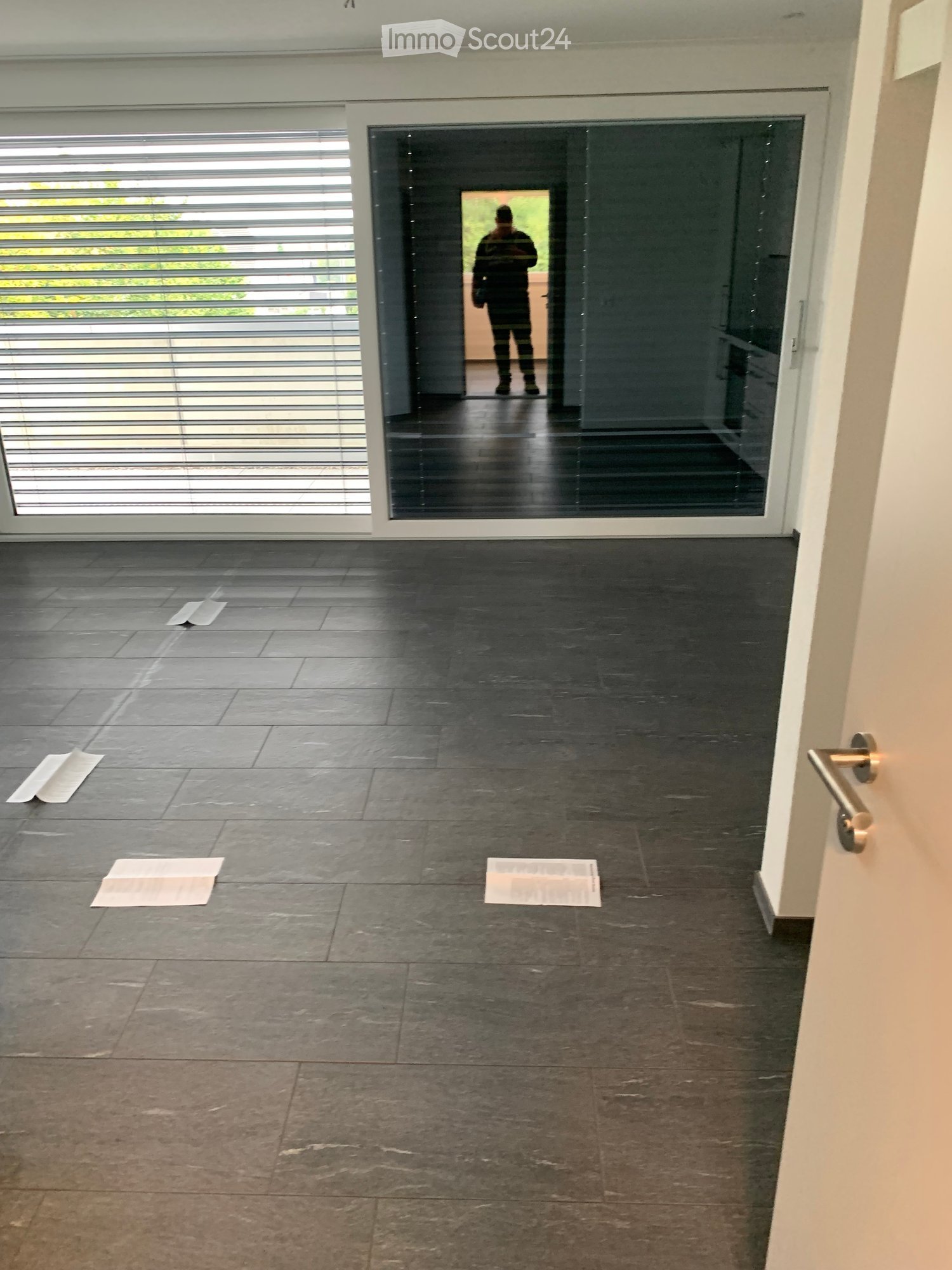 Empty room with dark tiled floor, white walls, large glass door with blinds, and a few scattered papers on the floor.