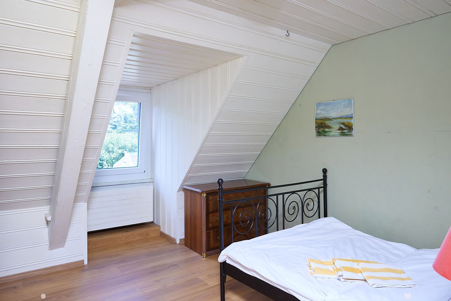 Cozy attic bedroom with wooden floors, slanted white ceiling, large window, bed with white linens, nightstand, and a small painting on the wall.