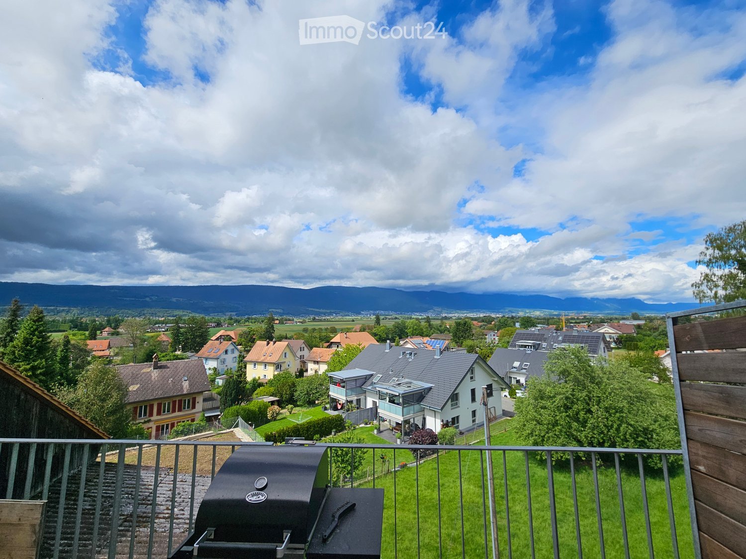 Balcony overlooking a suburban neighborhood with houses, green fields, and distant mountains under a partly cloudy sky. A grill is placed on the balcony.