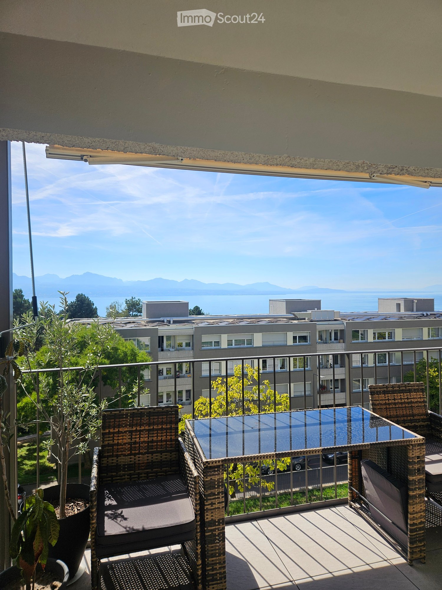Balcony with wicker chairs, a glass table, and a potted plant, overlooking a scenic view of a lake and mountains.