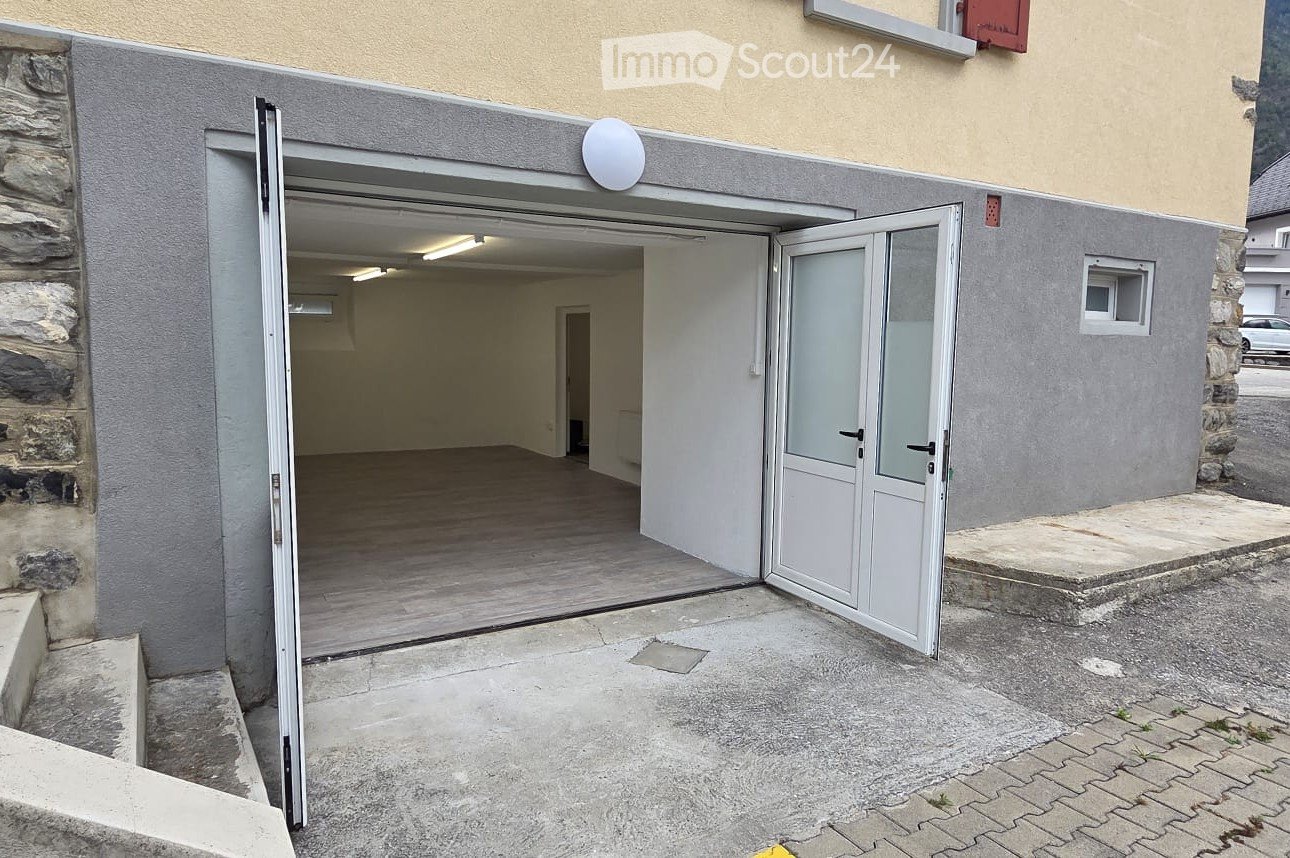 Open garage with wooden floor and white walls, adjacent to a stone building with a small window and a satellite dish.