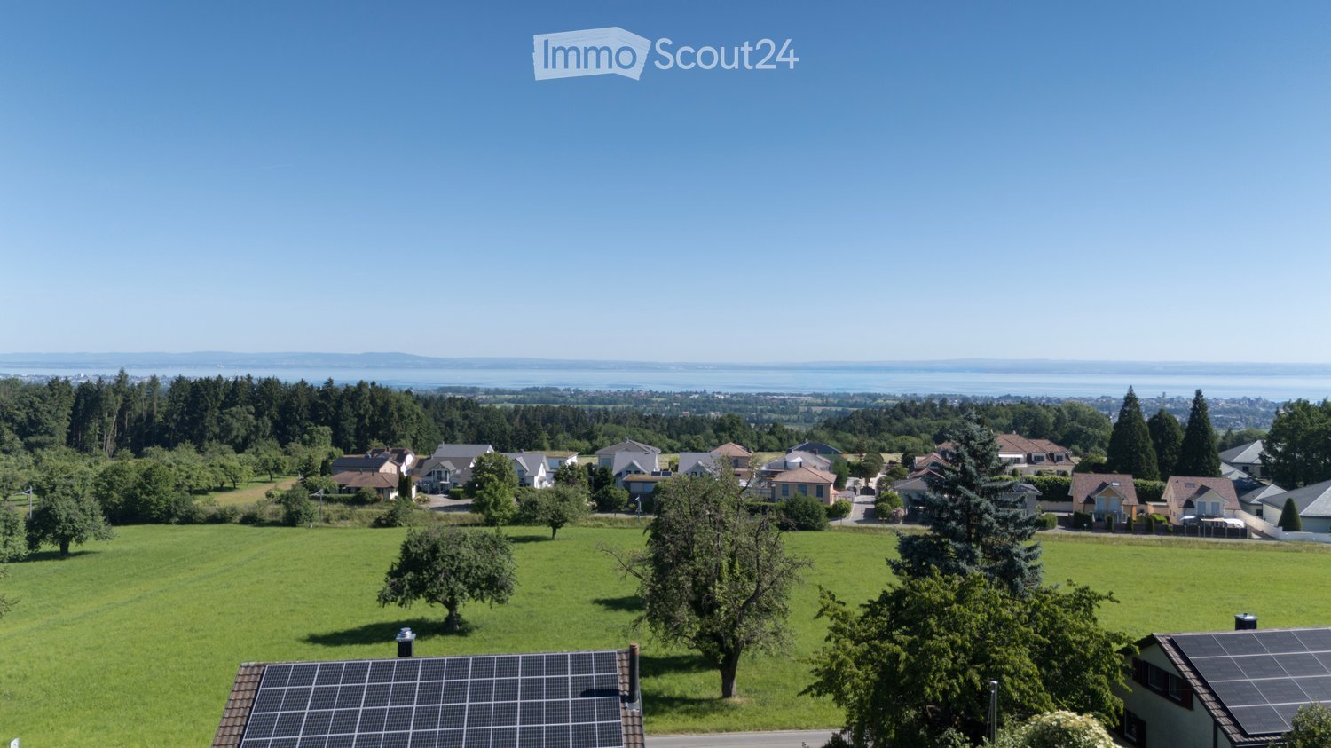 A panoramic view of a suburban neighborhood with houses, green lawns, trees, and a distant body of water under a clear blue sky.