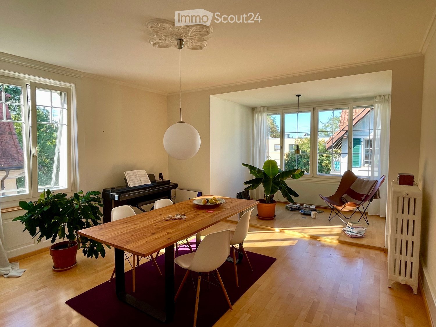 Bright, open room with wooden flooring, large windows, a wooden dining table with chairs, a piano, and potted plants.