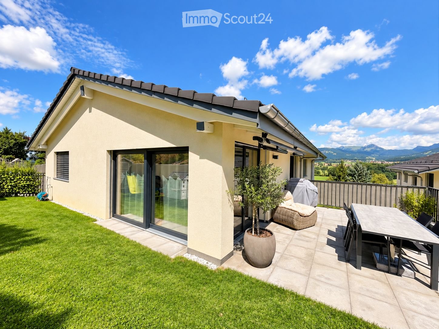Modern house with a well-maintained garden and a patio area featuring a table, chairs, a wicker sofa, and a potted plant. The house has large glass doors and a clean, contemporary design.
