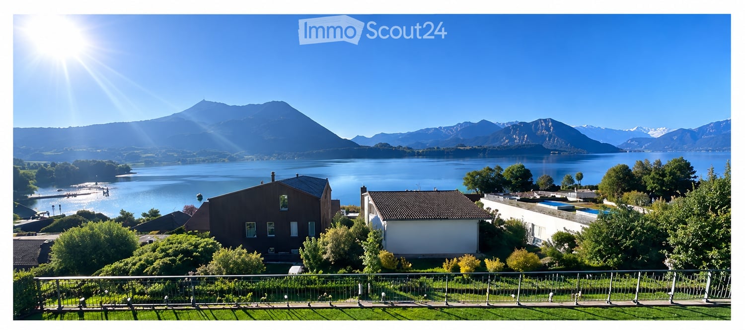 A scenic outdoor view featuring a lake, mountains, and a house with a balcony overlooking the landscape.