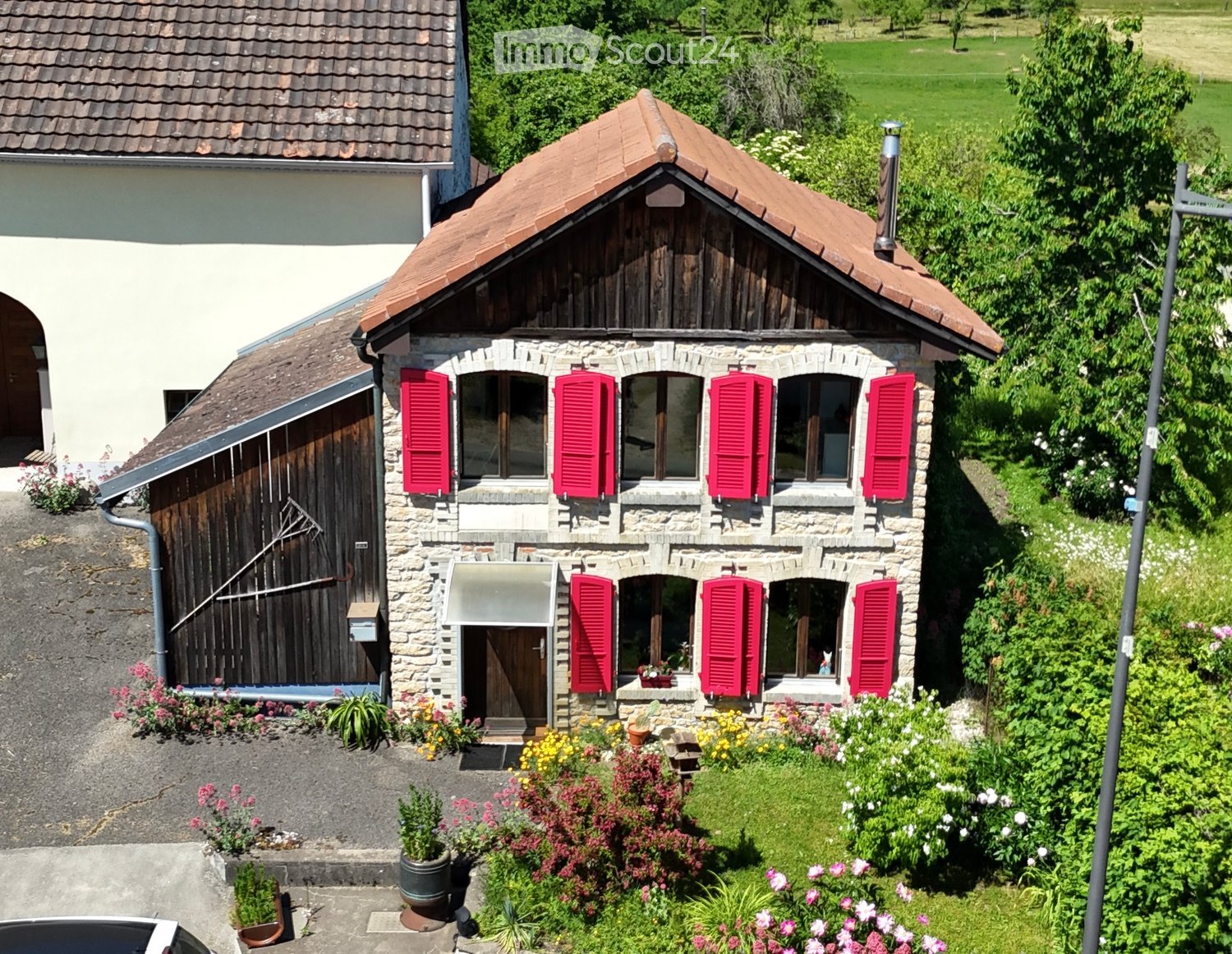 A charming stone house with a red-tiled roof, red shutters, and a well-maintained garden with colorful flowers and potted plants.